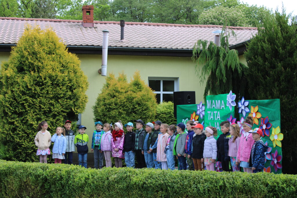 Występ dzieci z grupy najstarszej. Spiewają piosenkę i recytują wiersz dla Mamy i Taty. Uroczystość ma miejsce w przedszkolnym ogrodzie.