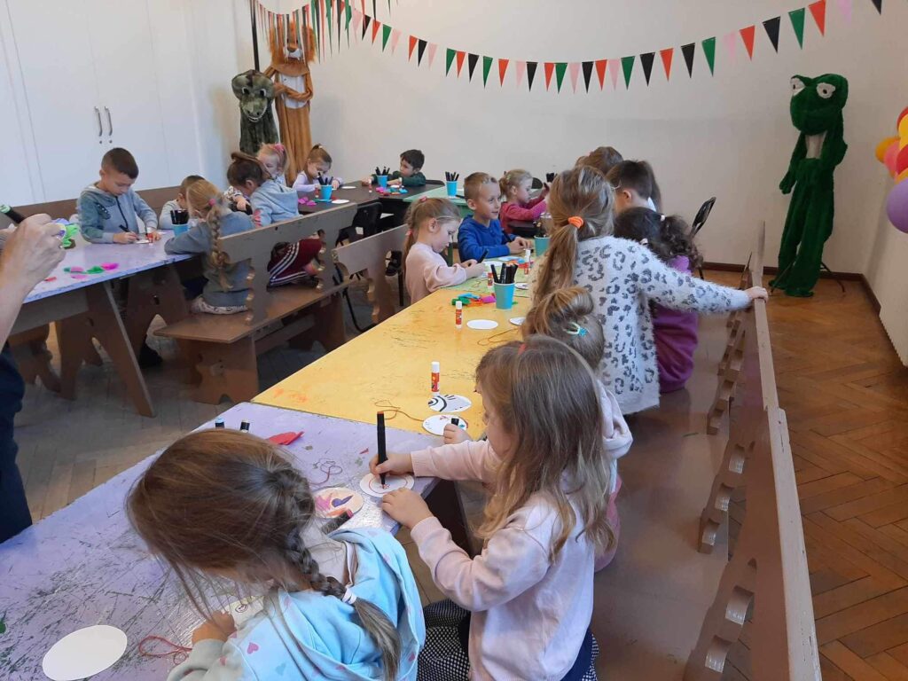 Dzieci siedzą przy stolikach w sali warsztatowj SDK. Biorą udział w warsztatach plastycznych i wykonują medalion.