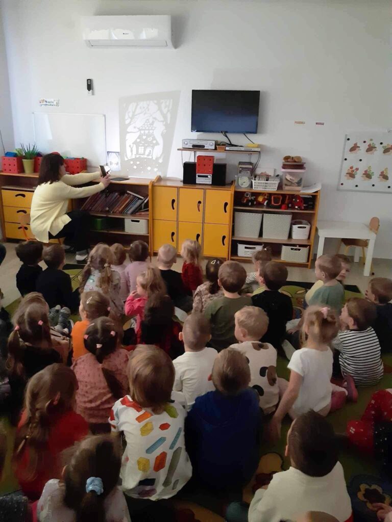 Teatrzyk ciei w sali przedszkolnej z okazji Andrzejek. Wszystkie dzieci siedza na dywanie obserwują obrazy prezentowane na ścianie do bajki JAŚ I MAŁGOSIA i słuchają czytanej bajki