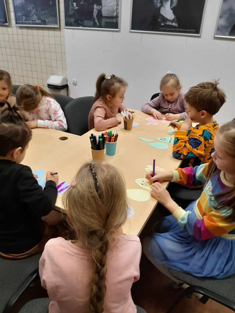 Dzieci rysuja zajączka i na warsztatach wykonuja ozdobe na patyczku. Pracują przy stołach w Domu Kultury.