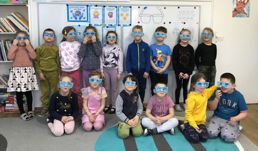 dzieci stoja pod tablicą , założyły na nos niebieskie okulary żeby uczcicć obchody Światowego Dnia Świadomości Autyzmu. Na zajęciach rozmawaiły o tym zaburzeniu