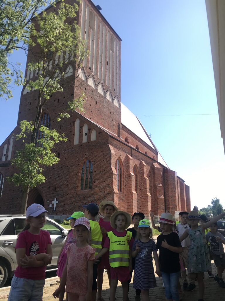 Przedszkolaki stoją przed dużym Kościołem w Sławnie, oglądają zabytek