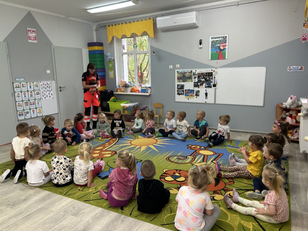 przedszkolaki siedza na dywanie , uczestnicza w spotkaniu z pania ratownik, ktora prezentuje maskotke misia ratownika