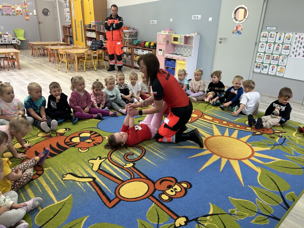 Pani ratownik demonstruje jak pomóc innej osobie, przedszkolaki siedzą w kole , jedna z dziewczynek odgrywa rolę osoby nieprzytomniej 