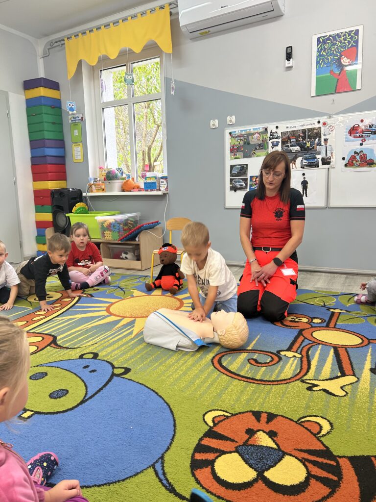 Chłopiec próbuje na fantomie wykonać prawidłowo uciski na klatce piersiowej, pozostałe dzieci obserwują kolege i słuchaja pani ratownik ktora opowiada o ratowaniu życia .