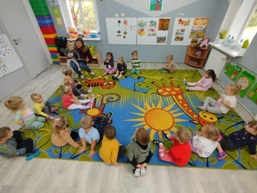 Dzieci siedza w sali przedszkolnej na dywanie, słuchają czytanych przez Panią Bibliotekarkkę wierszyków z okazji Dnia Głośnego Czytania