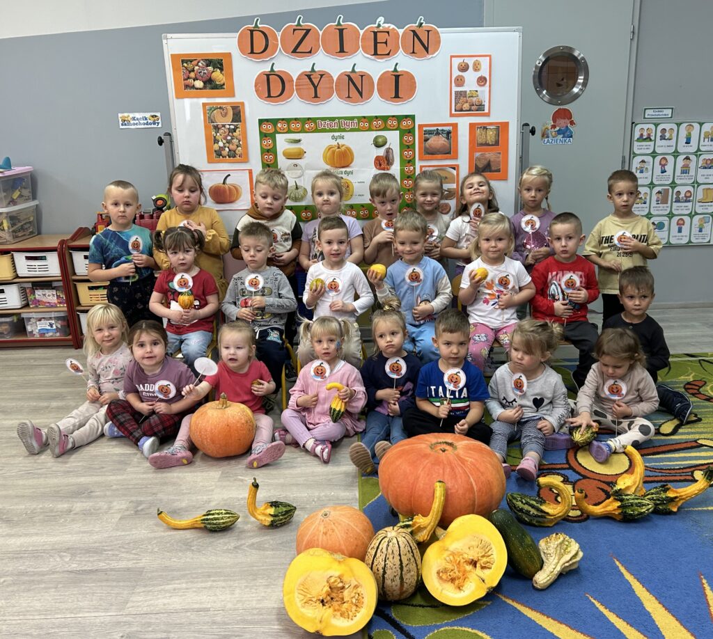 Dzieci w przedszkolu siedzą przed tablicą informacjną, na ktorej znajdują się informacje o obchodach DNIA DYNI. przed dzieć mi leżą prawdziwe okazy tego warzywa, w rękach dzieci trzymają pamiątkowe dynie na patyku.