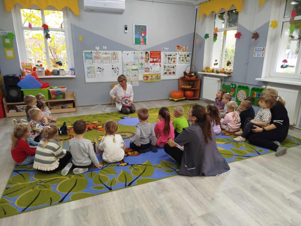 Wizyta pielęgniarki, która uczy Maluchy jak prawidłowo myć zęby. Dzieci siedzą na dywanie oglądają prawidłowe ruchy mycia zębów na specjalnym modelu