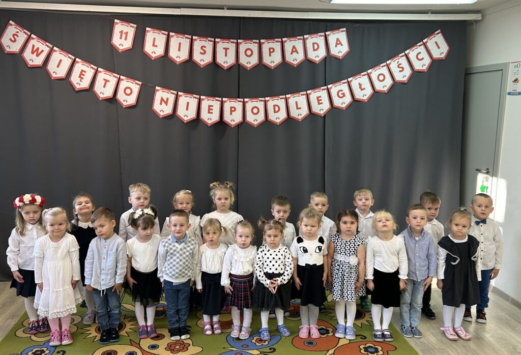 Grupa przedszkolaków stoi przed dekoracją związana z nadchodzącym świętem narodowym. Trzymają w reku Polska flagę . Przygotowuja sie do obchodów SWIĘTA NIEPODLEGŁOSCI. Wszystkie dzieci ubrane sa w strój galowy 