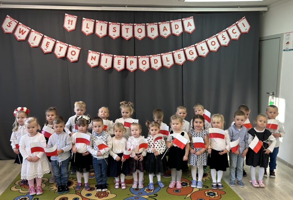 Dzieci w sali przedszkolnej stoją w dwuszeregu ubrane w strój galowy, przygotowują sie do nadchodzącego świeta Niepodległosci. Spiewaja hymn 