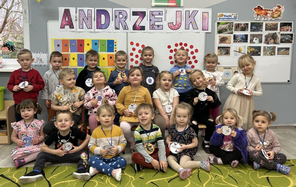 Dzieci stoją z andzrejkowymi symbolami na szyji przed tablicą informującą o dzisiejszym święcie w przedszkolu czyli ANDRZEJKACH 