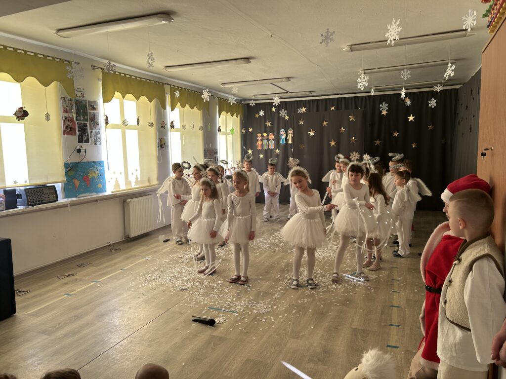 dziewczynki ubrane w stroje śnieżynek tańczą z magiczną śiecącą się różczką w rytm melodii świątecznej, pozostałe przedszkolaki oglądają występ