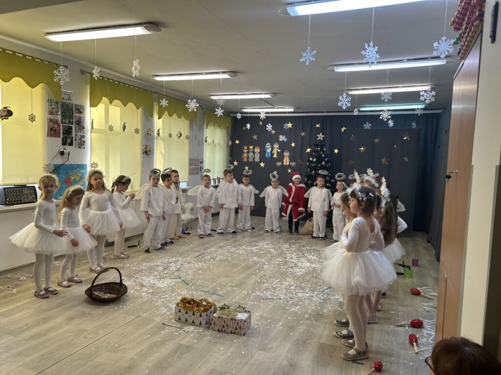 Przedszkolaki śpiewają kolędę ubrane w stroje do przedstawienia pt. W ANIOŁKOWIE, w tle stoi piekna choinka, którą przyniósł mikołaj. 