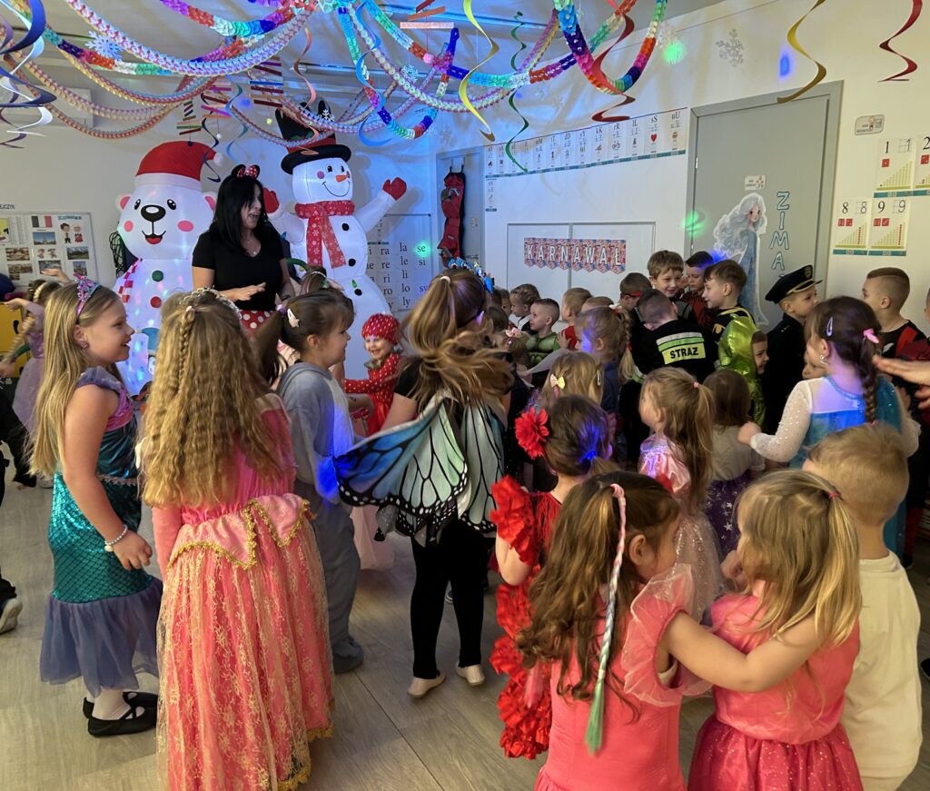 Bal karnawałowy w przedszkolu. Dzieci razem na sali przedszkolnej tańczą do muzyki. Wszytskie sa przebrane w stroje , pani wodzirej proponuje zabawy.