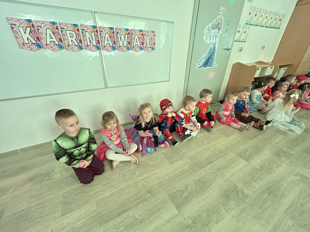 Dzieci siedza w sali przedszkolnej przebrane w stroje karnawalowe. oczekuja na swoja kolej i przeciąganie liny. Zdjęcie przedstawia bal karnawałowy .