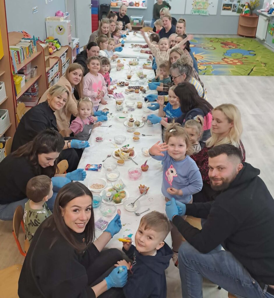 Na zdjeciu widac wszystkie dzieci z grupy I i ich rodziców. Siedza przy stole przy ozdobach  do babeczki wspolnie dekorują upieczone przez siebie słodkosci na kiermasz
