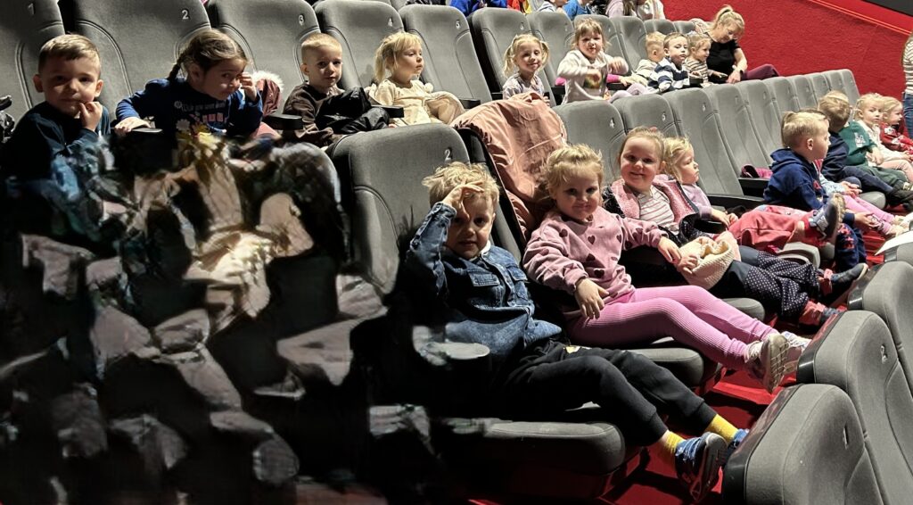 Dzieci z przedszkola siedza na fotelach w sdk i oczekuja na wystepy teatralne w ramach przeglądu. Kazde dziecko spogląda na aparat z usmiechnięta miną. 