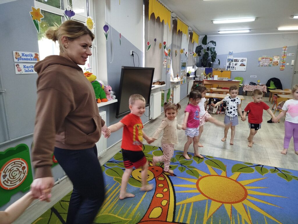 Dzieci uczestnicza w ćwiczeniach i zabawach ruchowych z zaproszoną trenerką fitness. Ubrane w stroje sportowe trzymają się za ręce i wykonuja okreslone ruchy. 