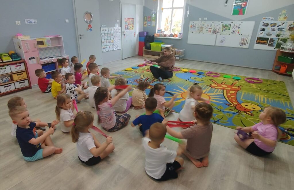 ćwiczenia z szarfami. Dzieci siedza na dywanie w siadzie skrzyżnym , wykouja ćwiczenia zgodnie z poleceniem trenera