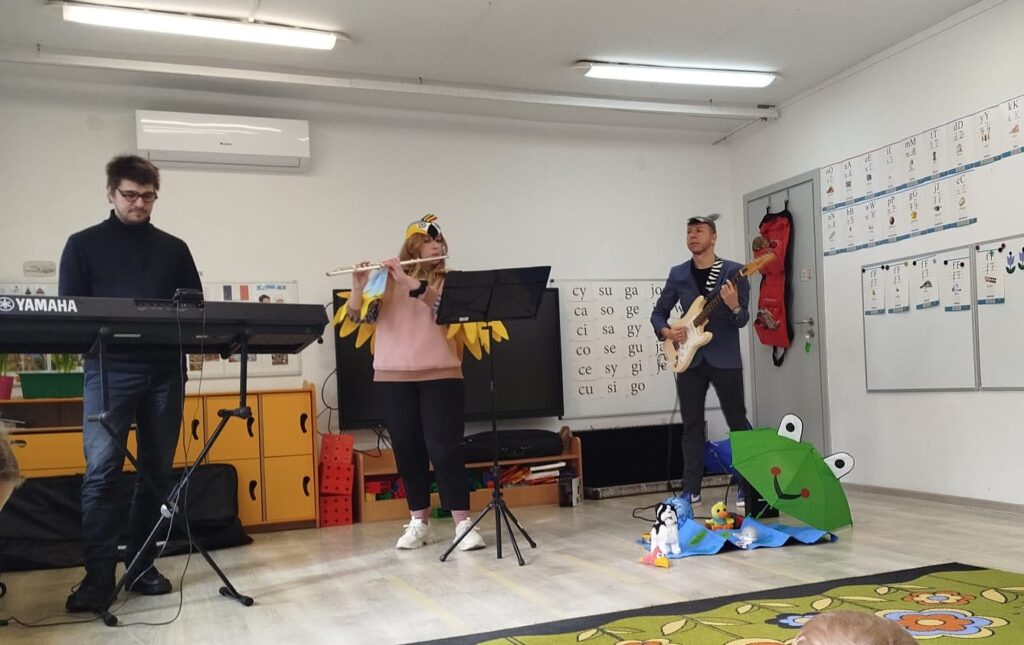 Muzycy FiLharmonii Koszalin przyjechali koncertowac w pzredszkolu. Zdjęcie przedstawia 3 muzyków grających na organach, flecie i gitarze. Dzieci słuchaja koncertu w ich wykonaniu