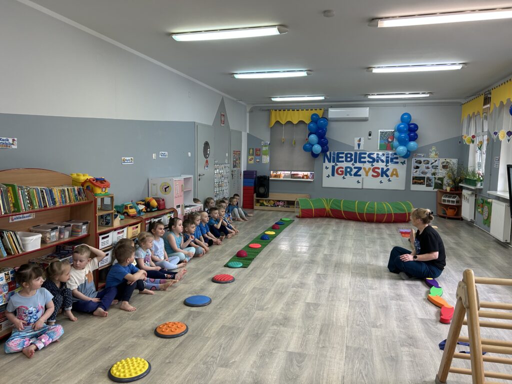 Przedszkolaki ubrane w niebieskie stroje sportowe siedza w rzędzie na sali gimnastycznej przygotowanej specjalnie na obchody NIEBIESKIECH IGRZYSK. Pani nauczycielka tłumaczy cel spotkania i planowanych zawodów spotrowych. Przed dziećmi ustawiony tor pzreszkod , ktory będa musiały pokonac 