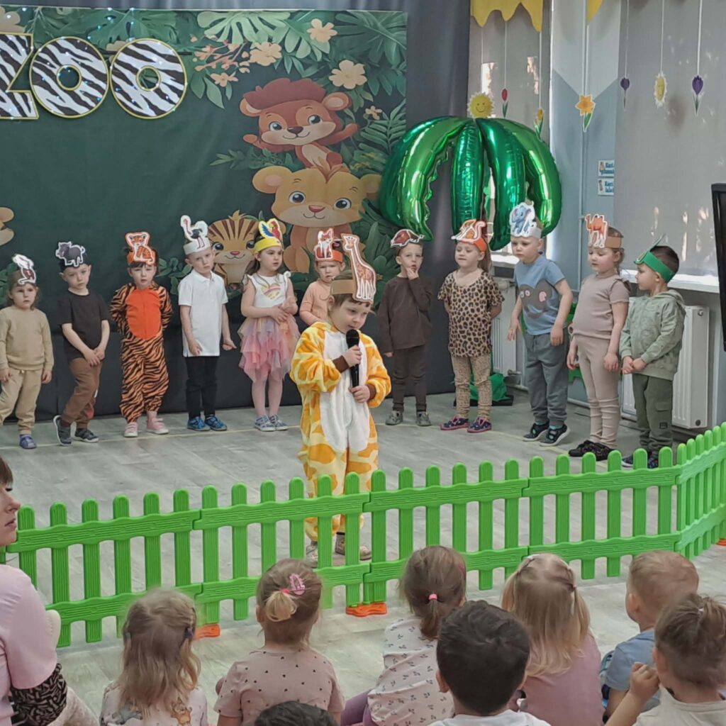 dZIEWCZYNKA UBRANA W STROJ ŻYRAFY RECYTUJE WIERSZYK, W TLE STOJA INNE PRZEDSZKOLAKI UBRANE W STROJE ZWIERZĄT. ODGRYWAJĄ PRZEDSTAWIENIE W RAMACH DNI TEATRU